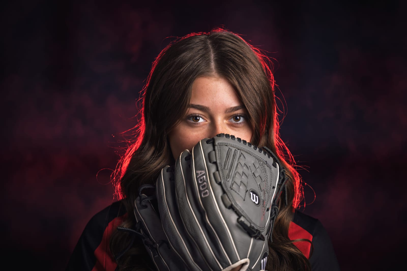 A softball player holding her glove across the lower half of her face under red rim light.