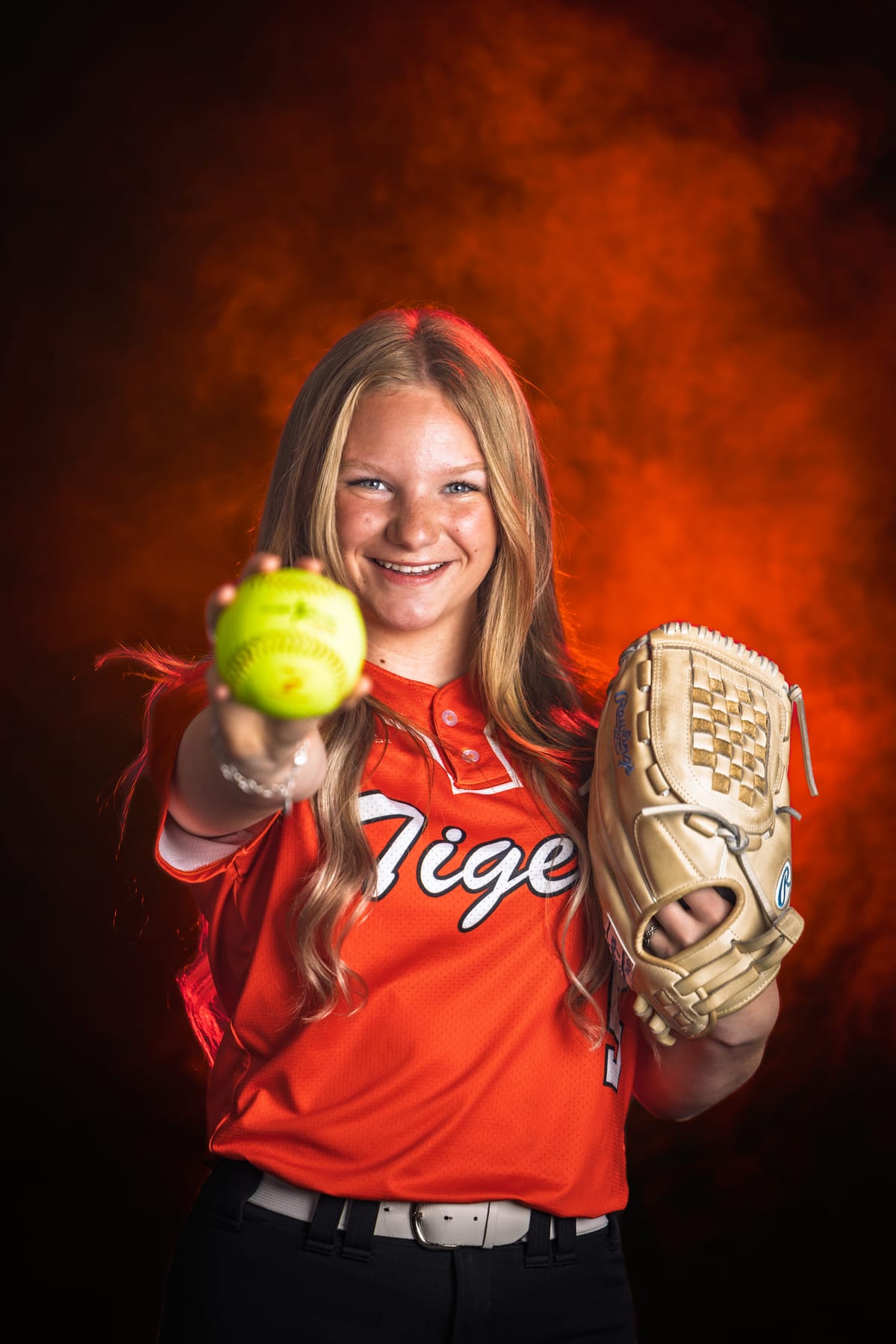 A softball player holding out a ball with her glove raised, red smoke behind.