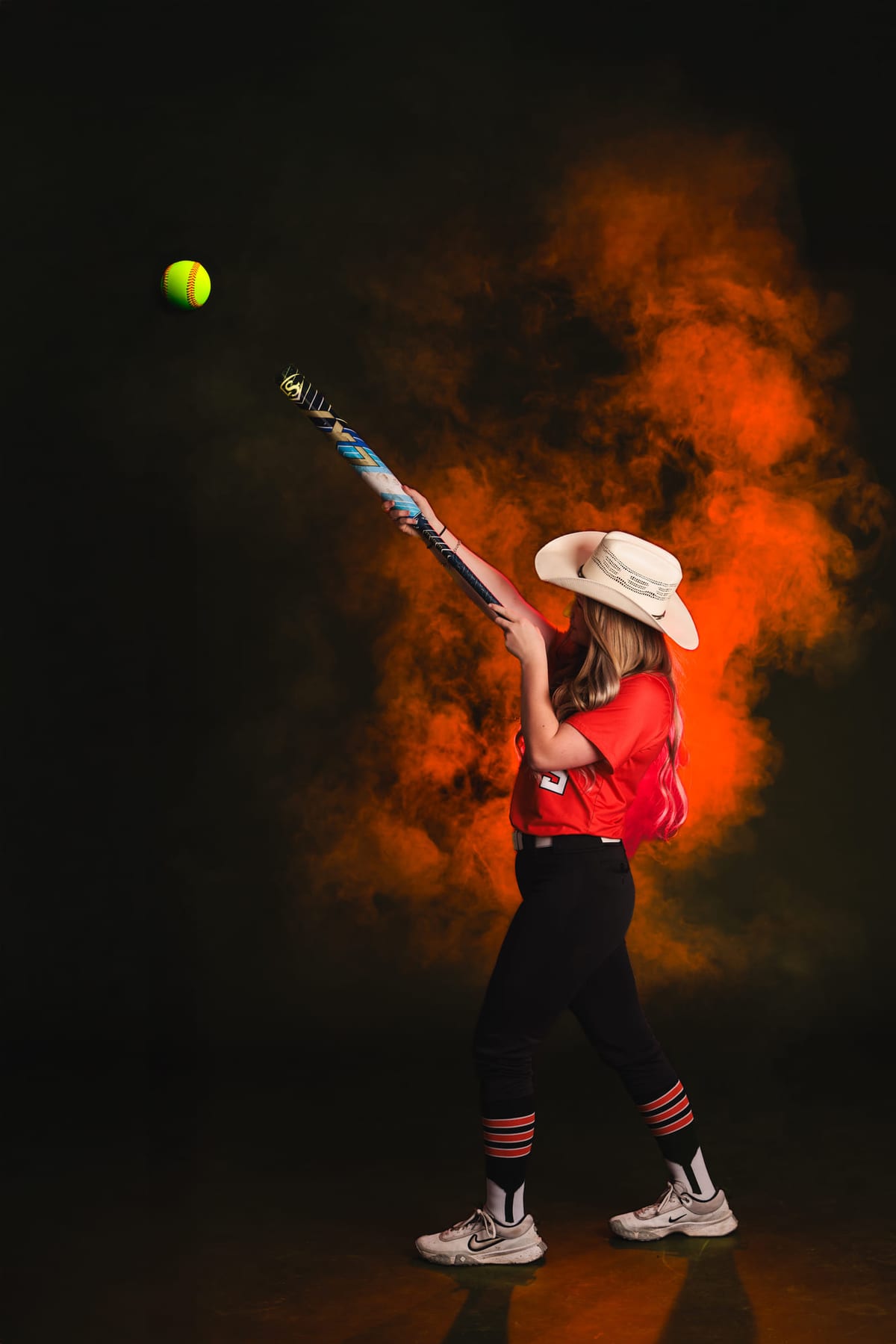 A softball player winding up to throw, red smoke rolling across a dark set.