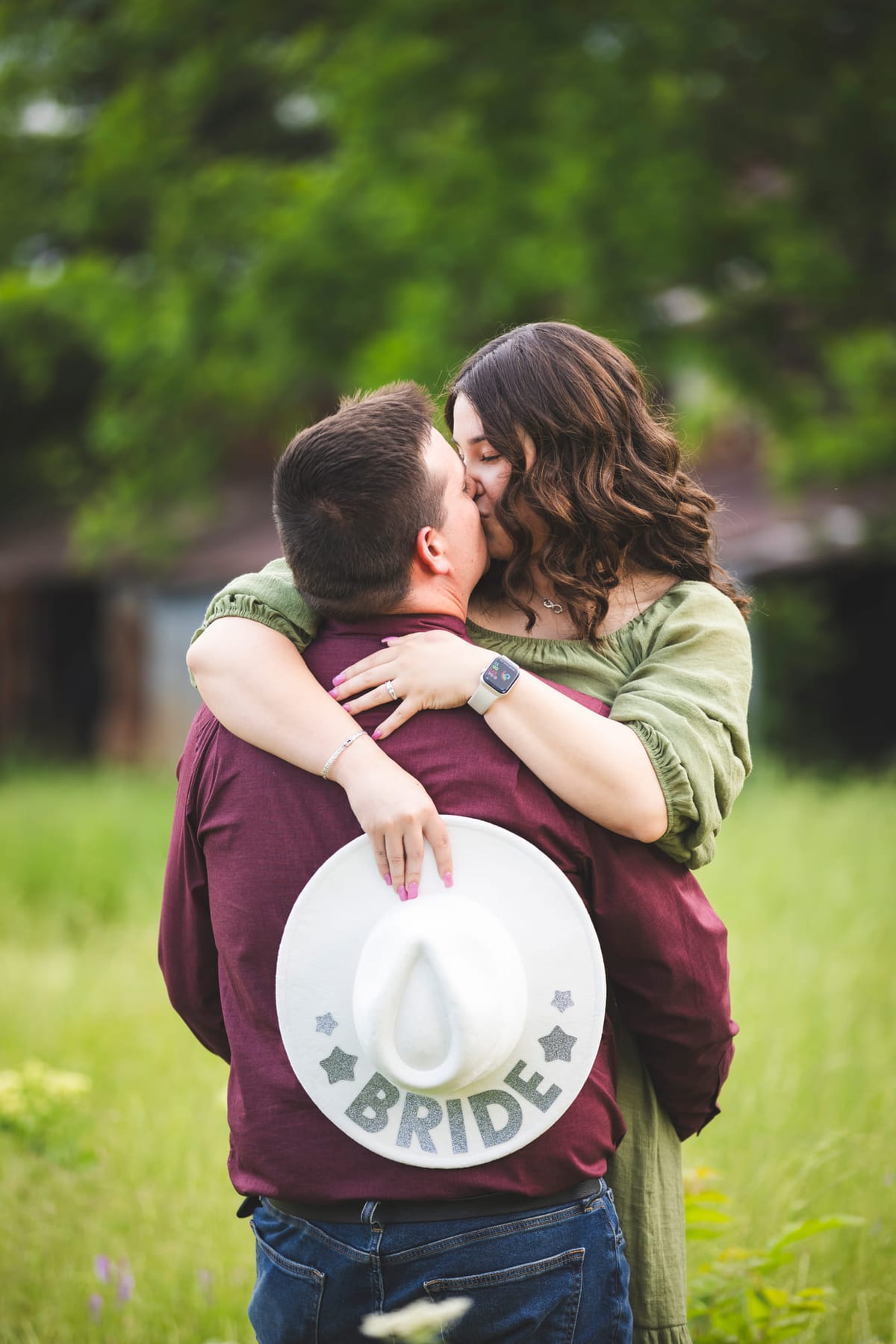 An engaged couple embracing in a field, a white hat lettered ‘BRIDE’ held behind his back.