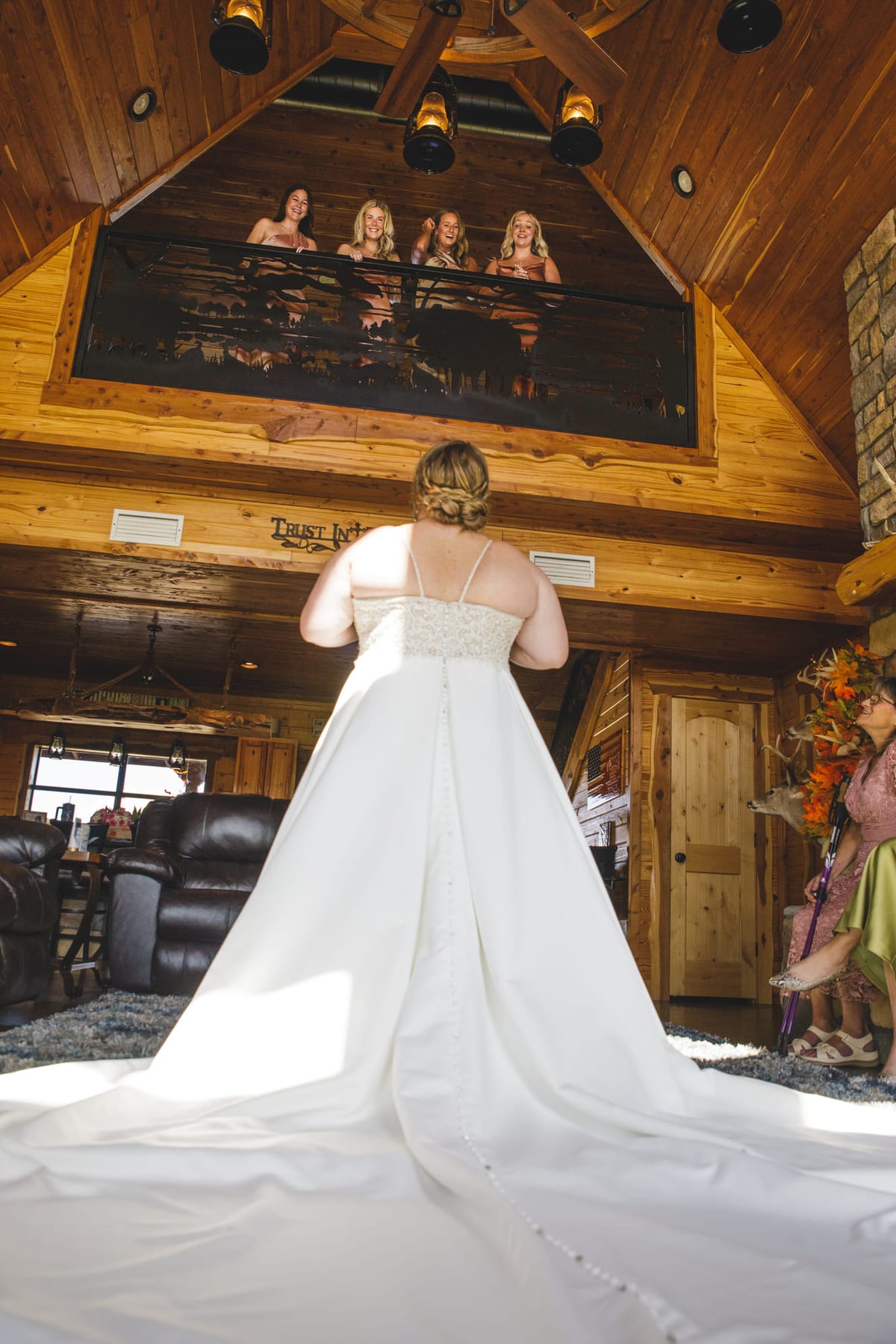 A bride seen from behind in a lodge hall, her long train laid out across the floorboards.