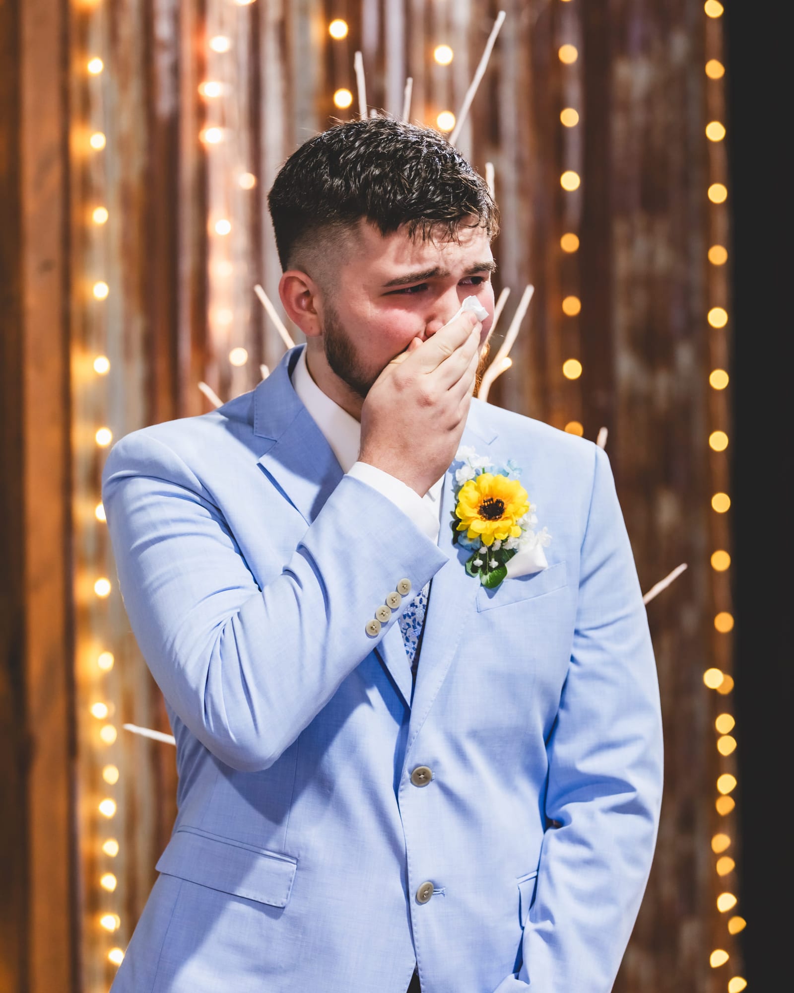 A groom in a pale blue suit with a sunflower boutonnière covering his mouth with one hand, strings of lights hanging behind him.