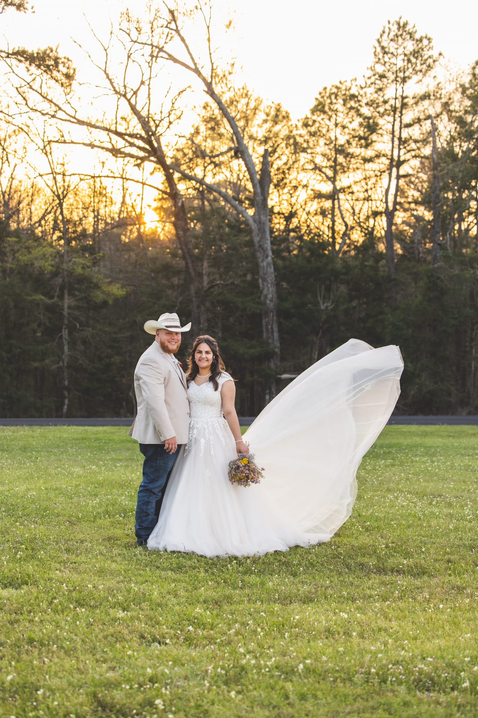 A couple standing in an open field at sunset, the bride’s train swept out wide across the grass.