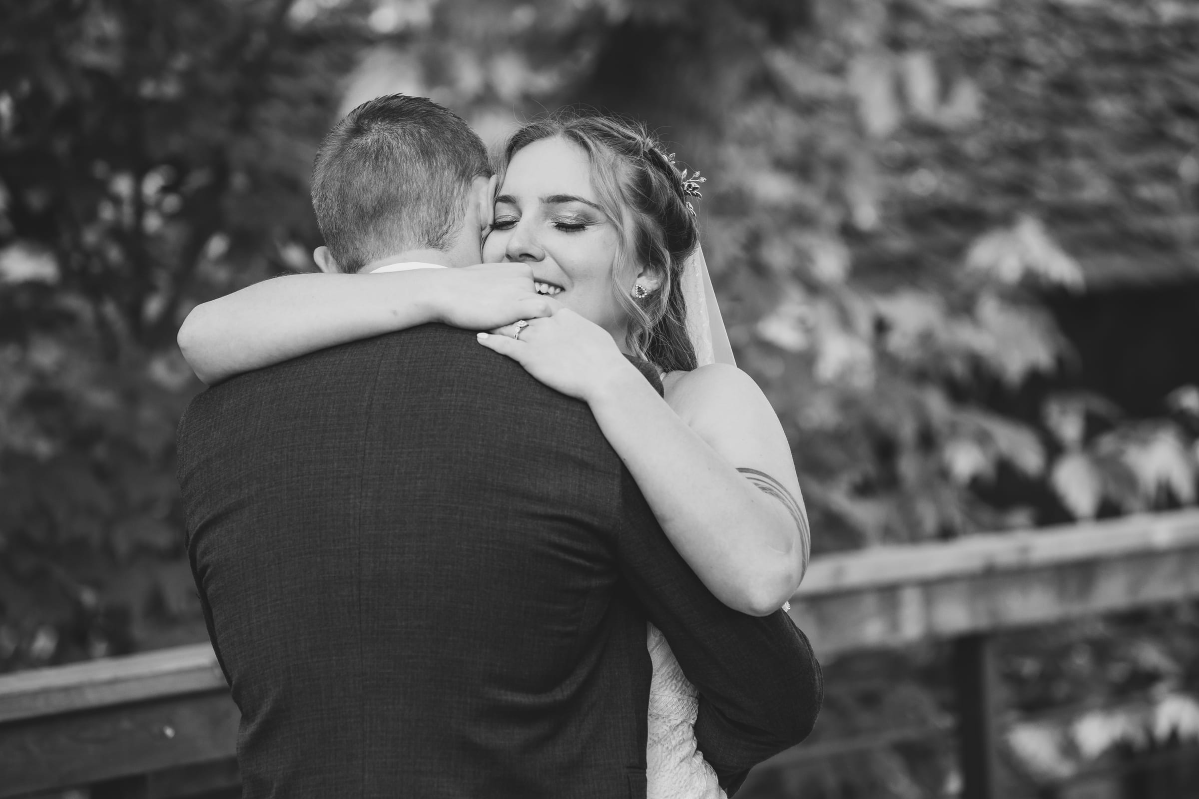 A bride with both arms around her groom’s neck, laughing, photographed in black and white.