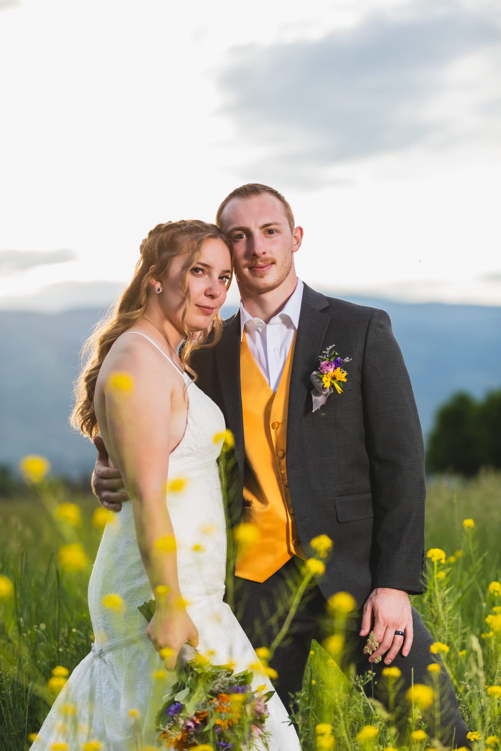 A couple standing together in a field of yellow wildflowers, the groom in a mustard tie and waistcoat.