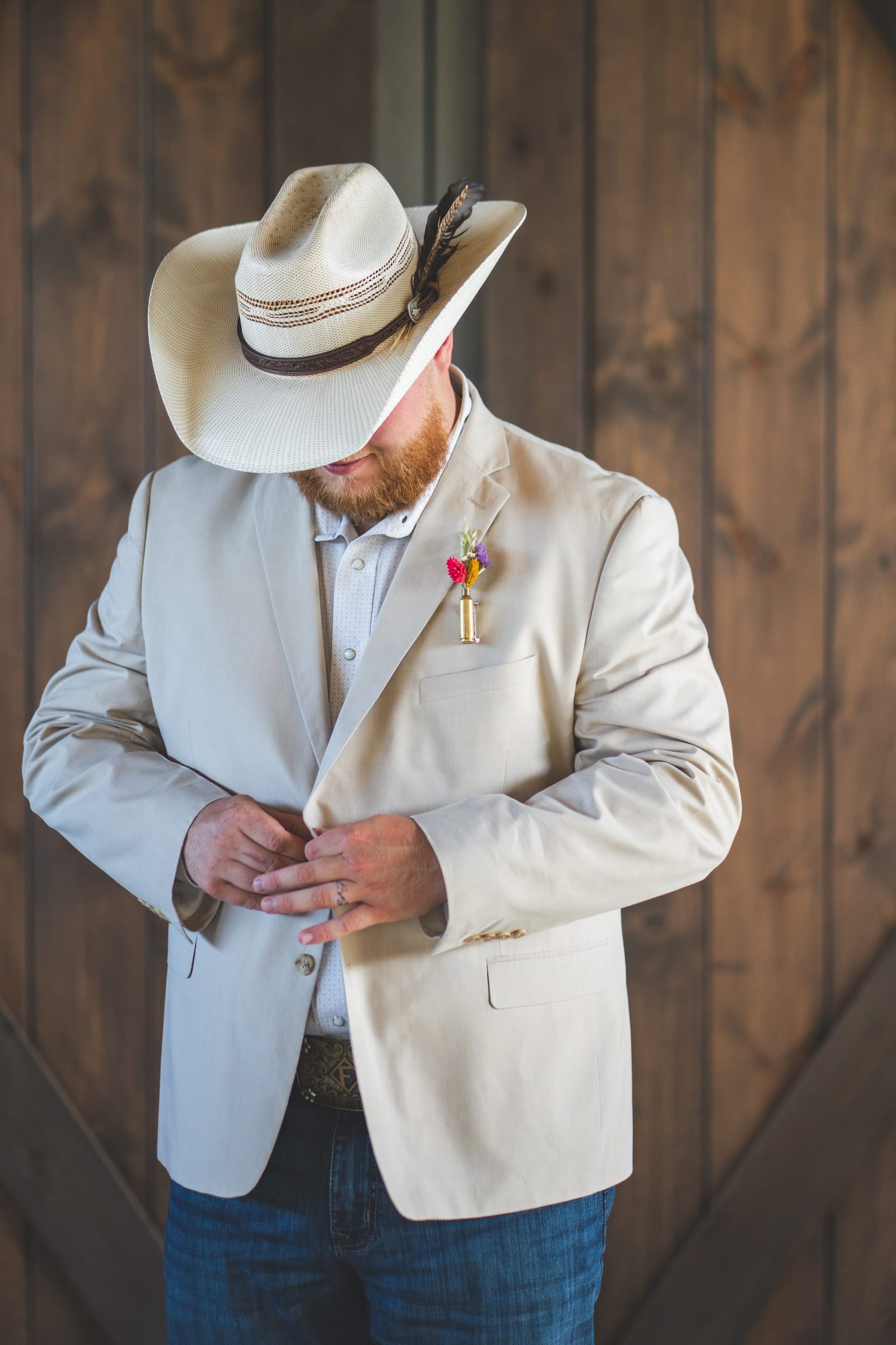 A groom in a cream jacket and felt hat fastening a button, head down.