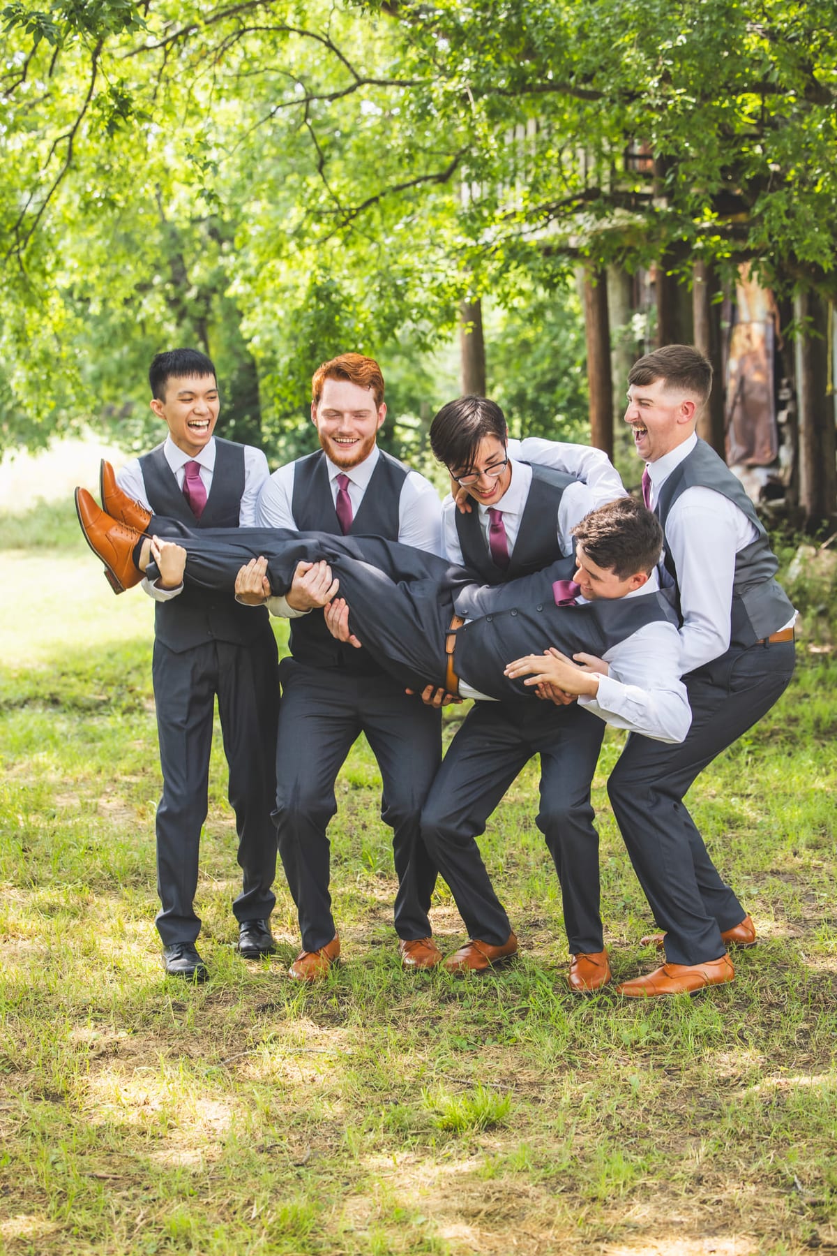 Groomsmen carrying the groom horizontally across the grass, all of them laughing.