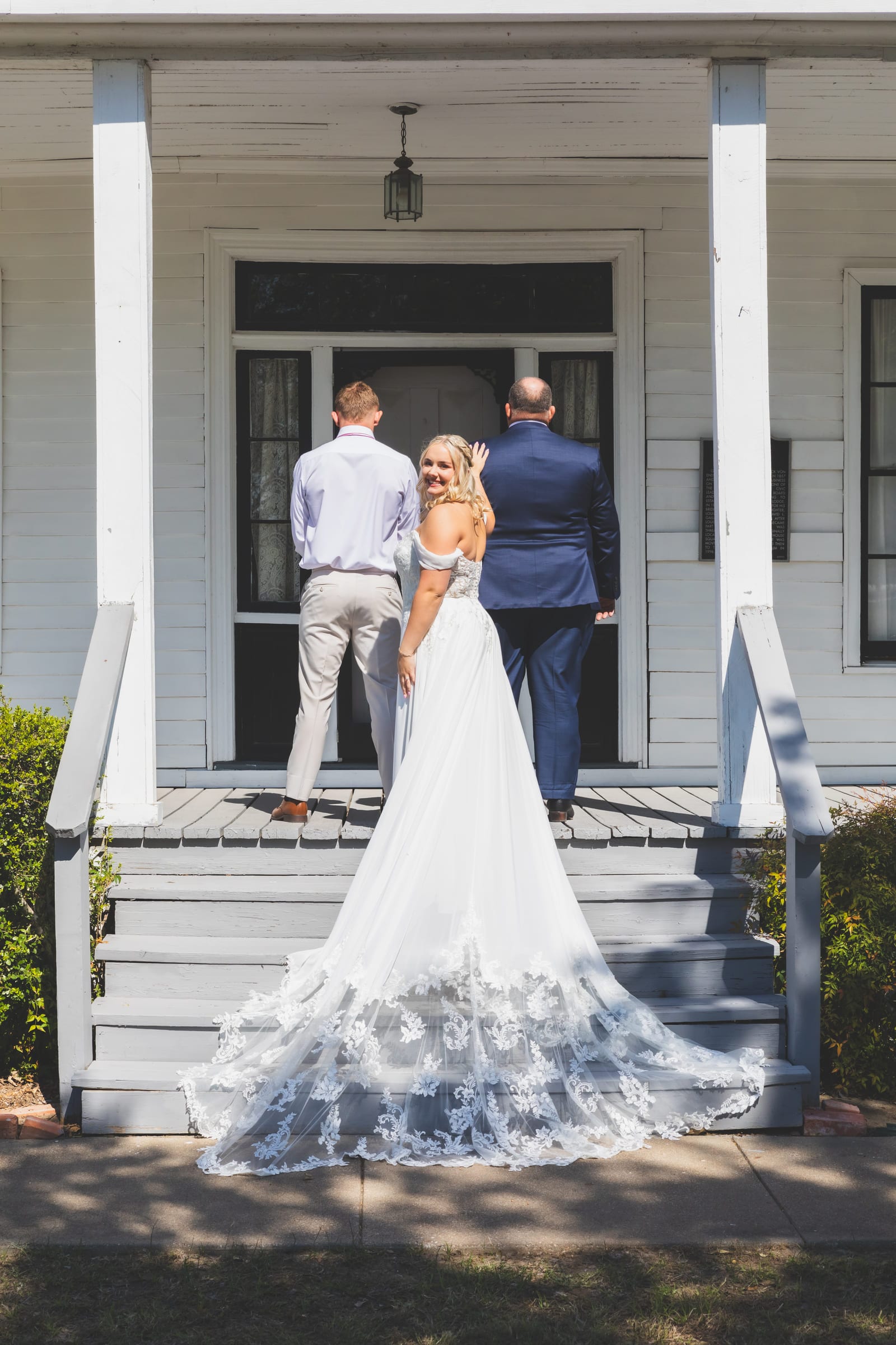 A couple embracing on the porch of a white clapboard house, the bride’s embroidered train spilling down the steps.