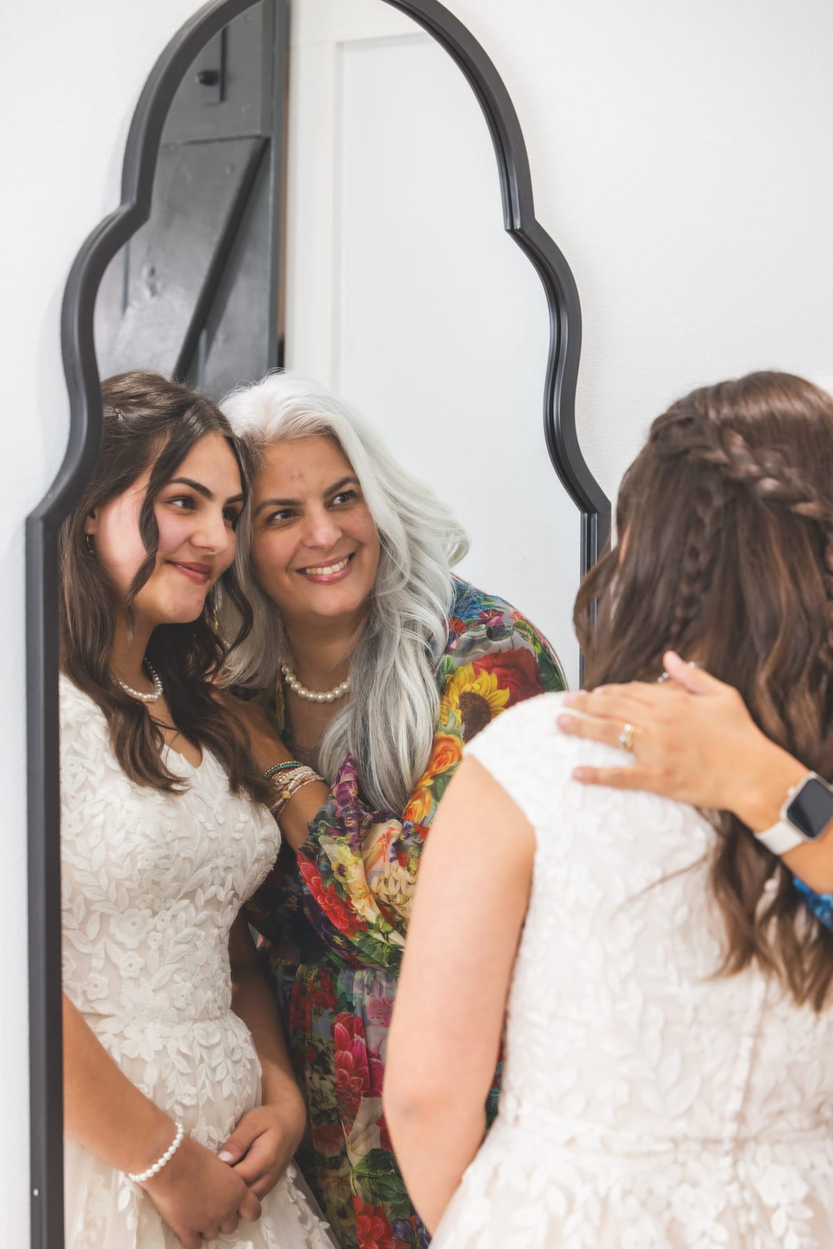 A bride reflected in an arched mirror while two others fasten her dress behind her.