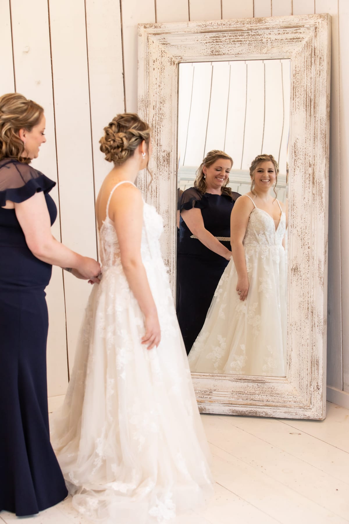 A bride at a full-length mirror with her attendants adjusting the gown behind her.