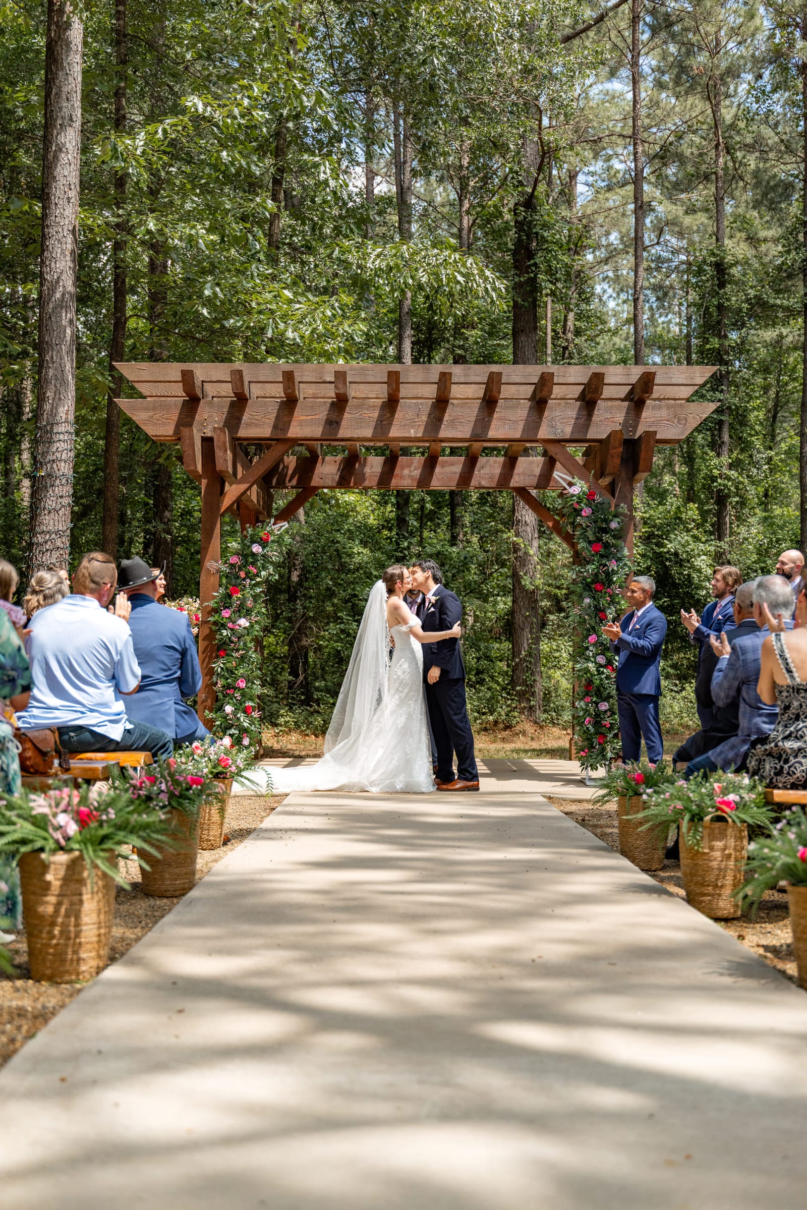A bride and groom kissing beneath a timber arbour at the end of a long aisle, guests standing on both sides.