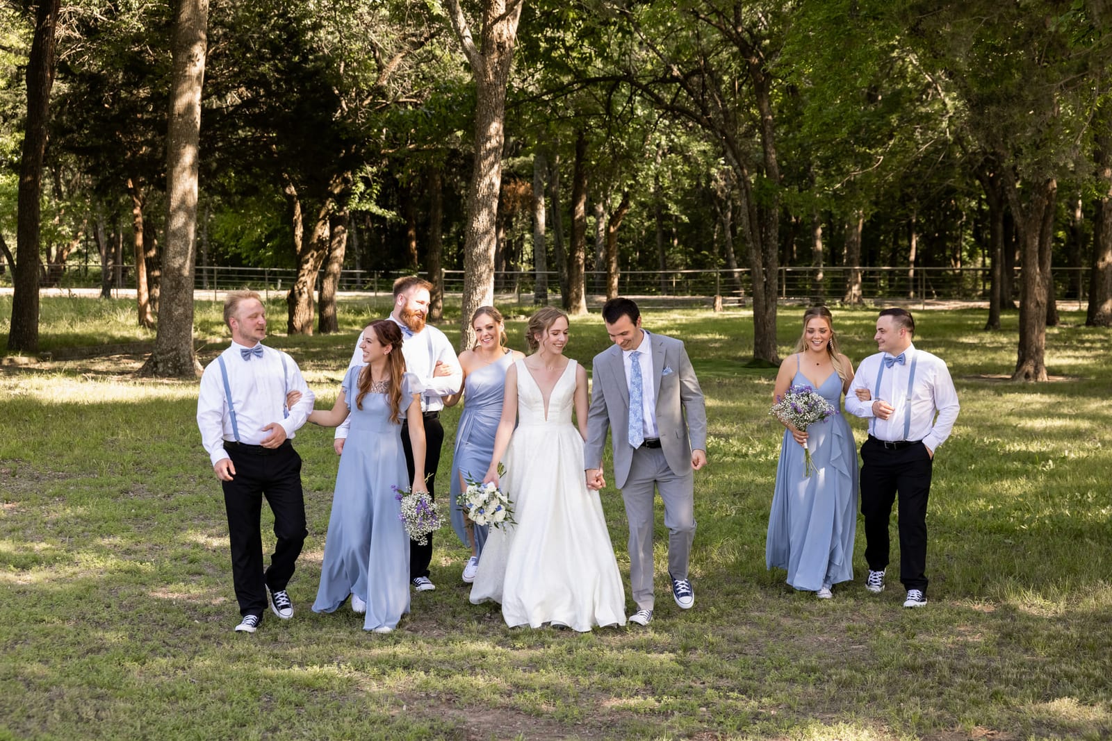 The full wedding party spread out under tall trees in dappled afternoon light.