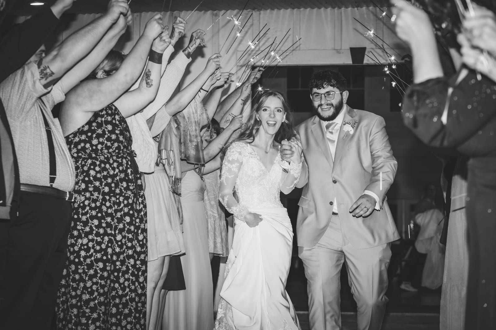 A black-and-white reception frame, guests crowding in around the couple with arms raised.