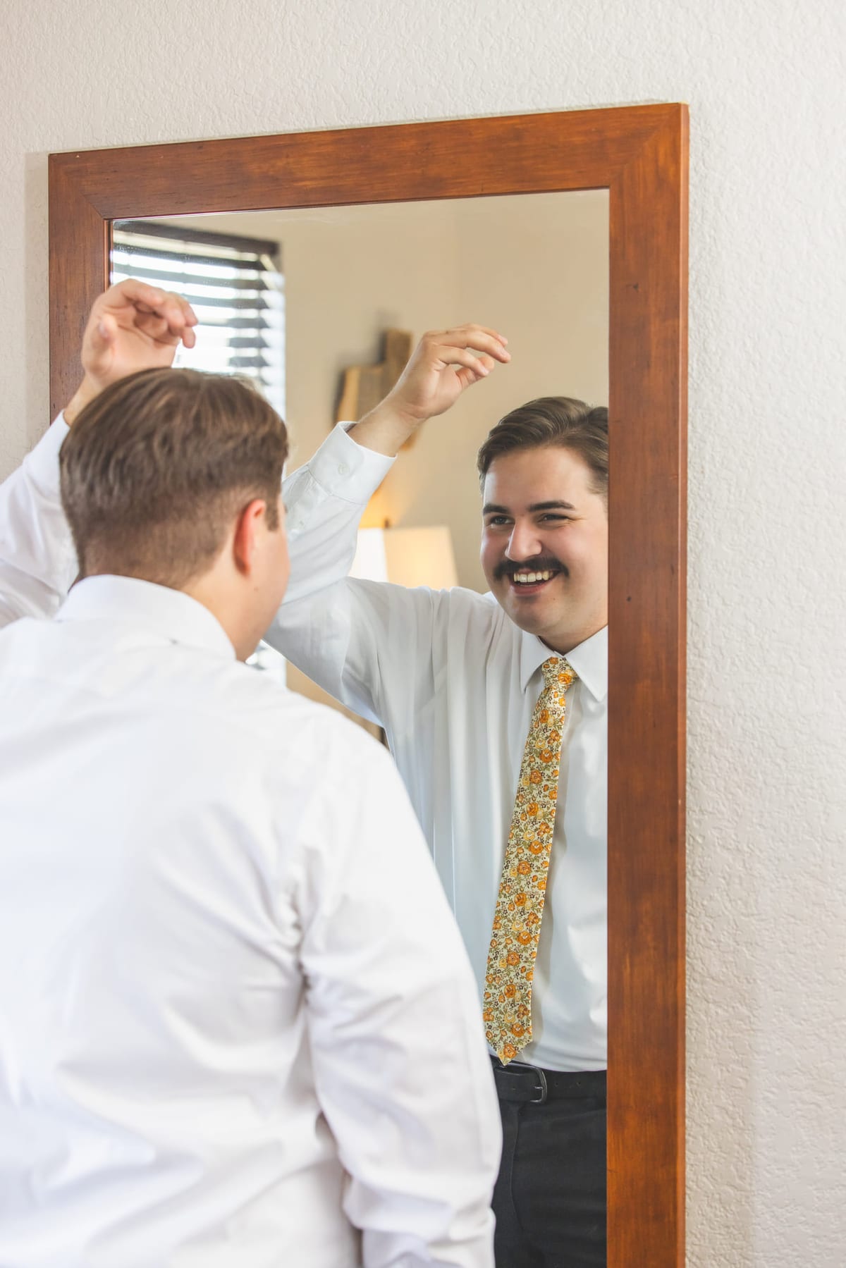 A groom having his tie straightened in front of a mirror, both men reflected in the glass.