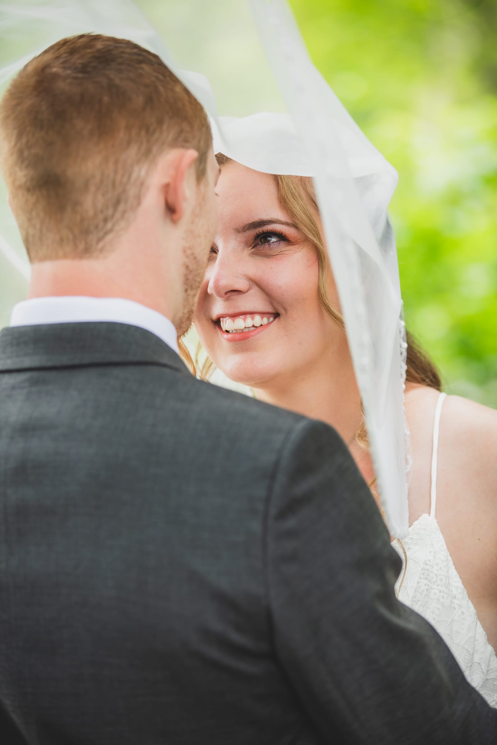 A bride and groom cheek to cheek beneath a lifted veil, her eyes to the camera.