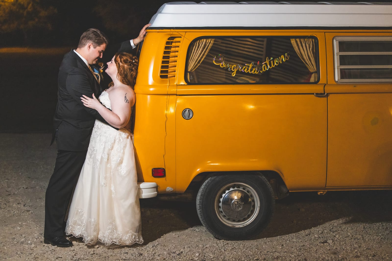 A bride and groom leaning together against an orange VW camper lettered ‘Congratulations’.